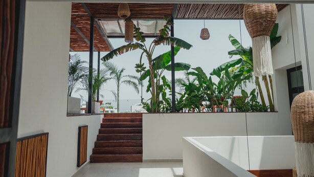 Un interior moderno con una escalera que lleva a un balcón con vistas a exuberante vegetación y al mar.