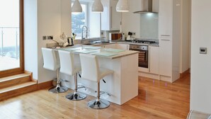 Een moderne keuken met een centraal eiland, witte barkrukken, roestvrijstalen apparatuur en een groot raam dat natuurlijk licht binnenlaat.