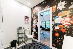 Une salle de bain avec des toilettes, un tapis rayé noir et blanc, une étagère en métal noir et une fresque florale sur le mur.