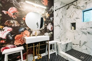 Une salle de bain avec un papier peint à motifs floraux, un lavabo blanc, des robinetteries dorées et un sol carrelé en noir et blanc.