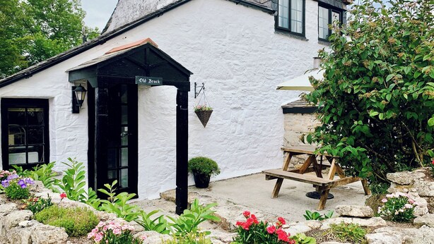 Um edifício branco com porta preta, um letreiro com a inscrição 'Old Brock' e um banco de pedra no jardim.