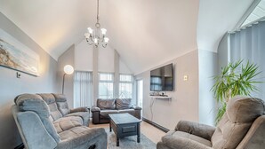 A spacious living room with a high ceiling, a chandelier, a flat-screen TV mounted on the wall, and a large window with blinds.