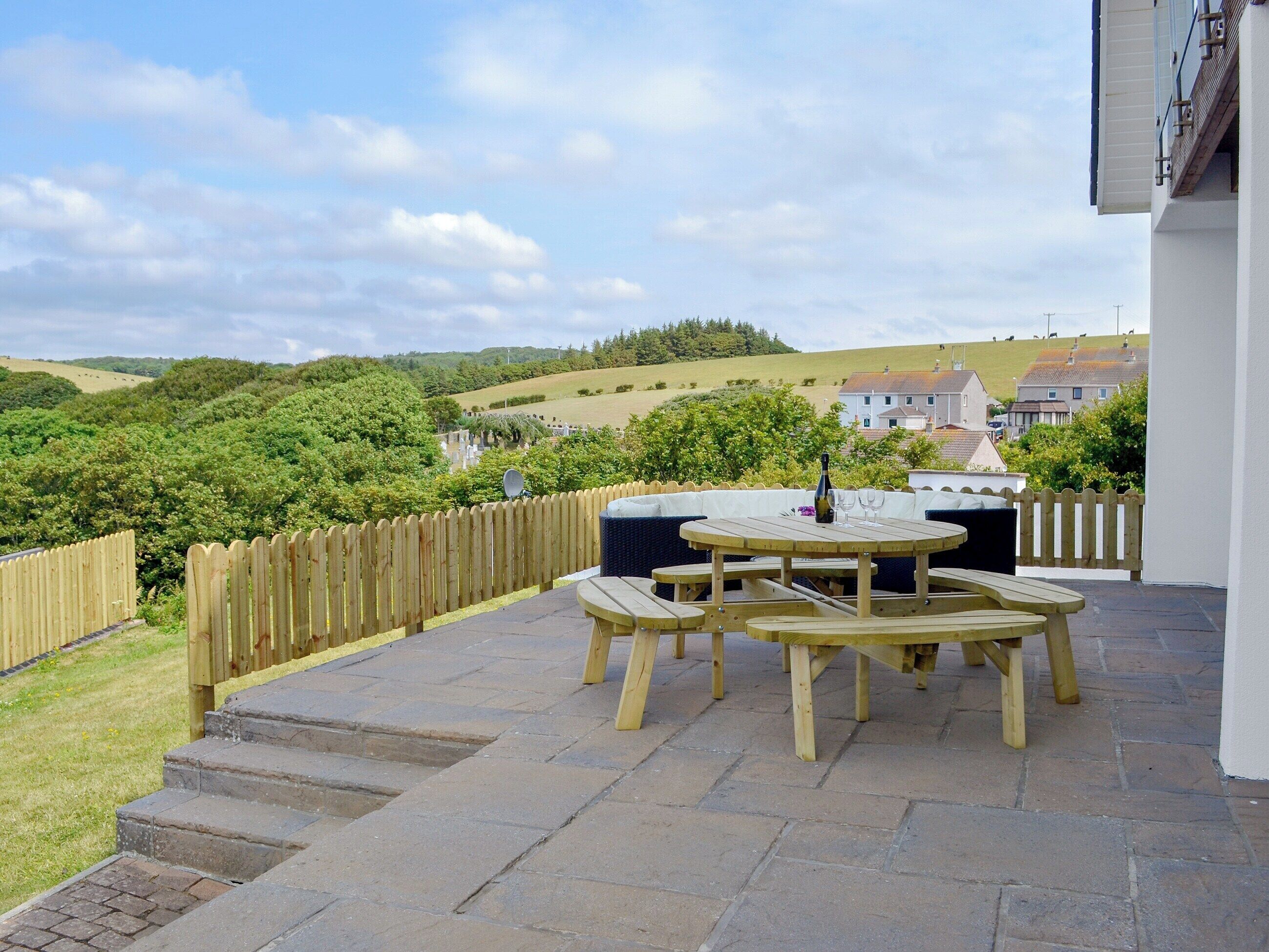 A patio with a wooden table and benches, a bottle of champagne, and a view of the countryside.