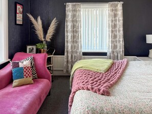 A bedroom with a pink sofa, a bed with a floral duvet, a radiator, and a window with floral curtains.