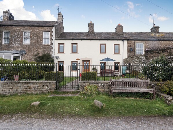A traditional stone house with a white facade, multiple windows, and a front garden with a bench.