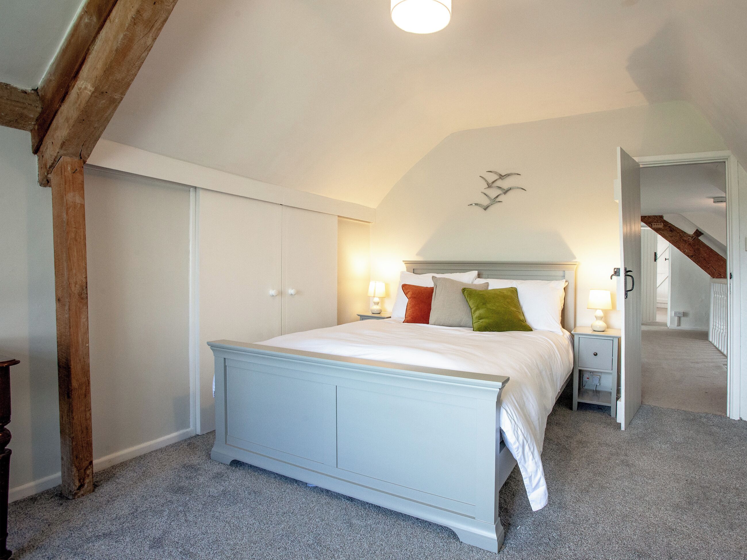 Een slaapkamer met een groot bed, houten balken, een schuin plafond en een trap naar een andere kamer.