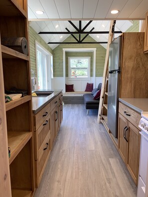 A compact kitchen with wooden cabinets, a built-in bench, and a window overlooking greenery. - Tiny House at Bidwell Canyon Farm (Fort Bidwell)