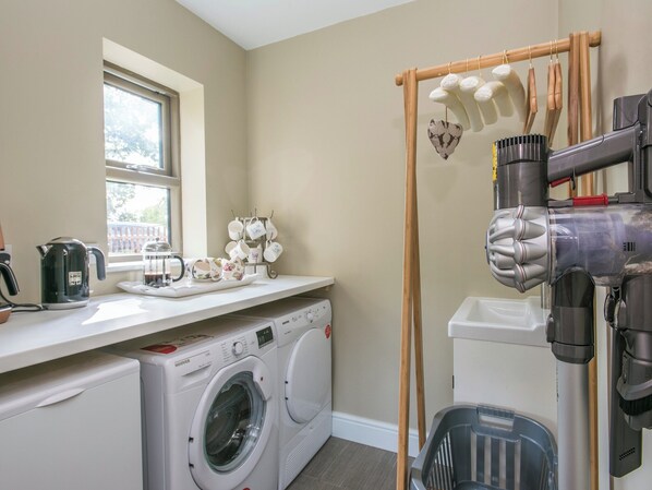 A laundry room with a front-loading washer and dryer, a window, a drying rack with clothes, and a vacuum cleaner.