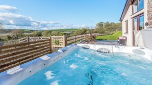 Un jacuzzi con cubierta de madera, vista a un paisaje rural y un edificio con grandes ventanales.
