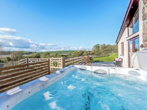 A hot tub with a wooden deck, a view of a rural landscape, and a building with large windows.