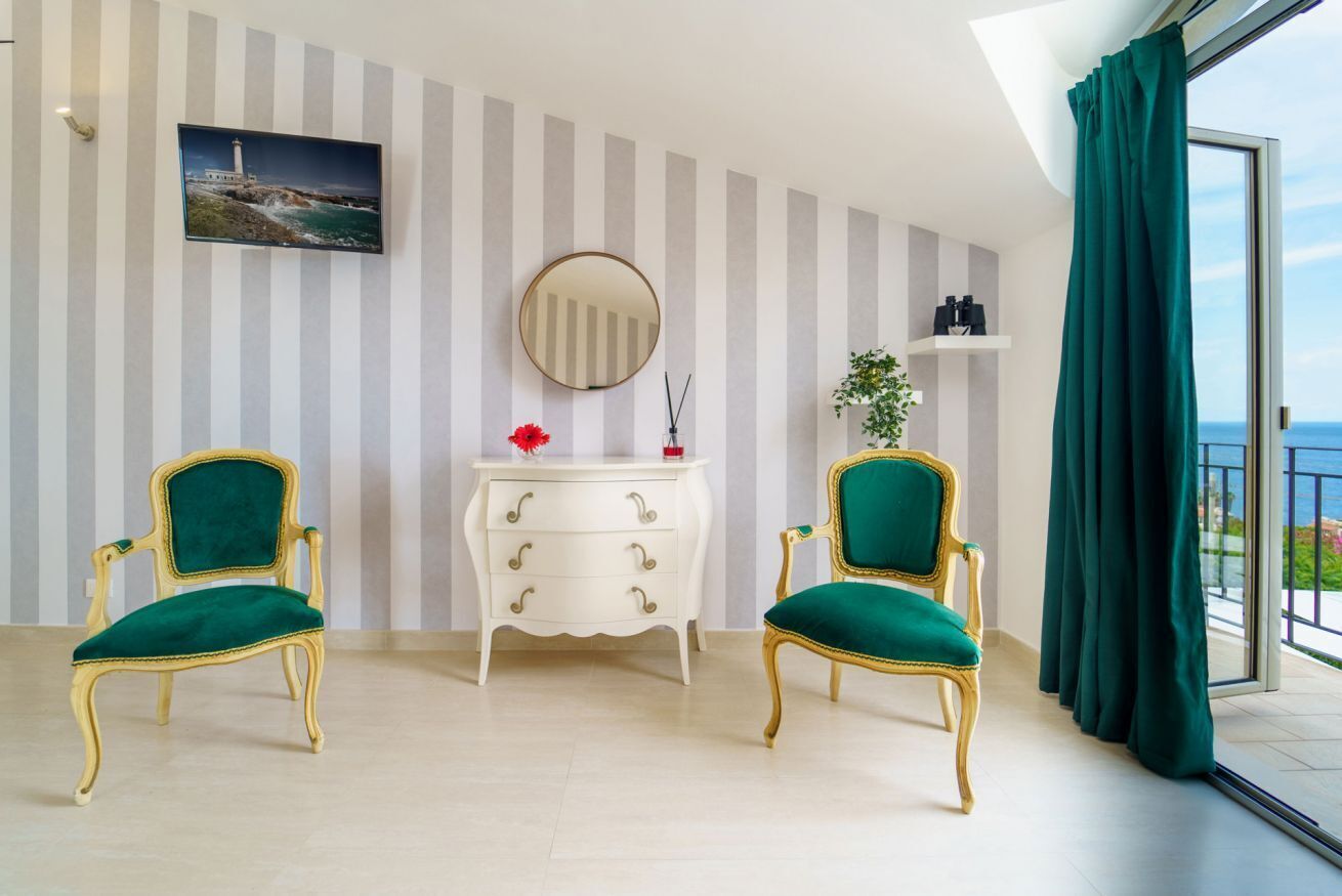 Een kamer met twee groene bekleden stoelen, een witte dressoir met lades, een ronde spiegel, een plank met flessen en een schilderij van een vuurtoren aan de muur.