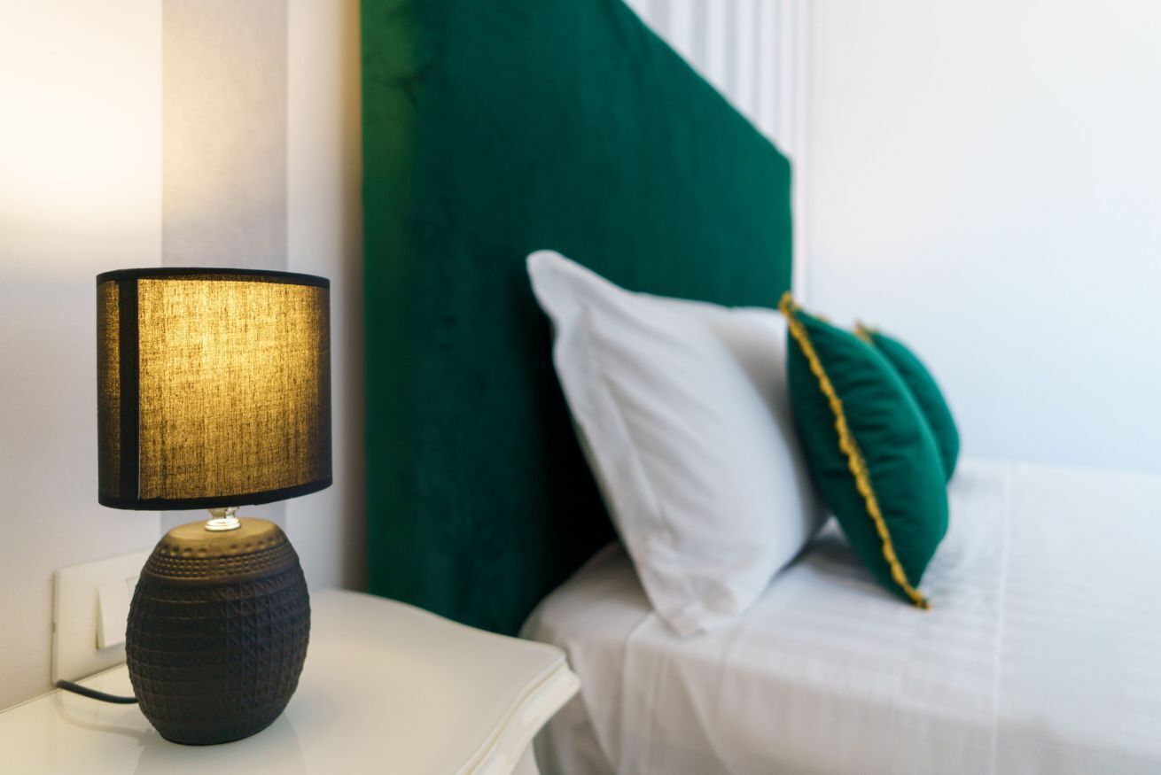 Een bed met witte lakens en groene kussens, een lamp met een gestructureerde kap op een witte nachtkastje, en een groen hoofdeinde.