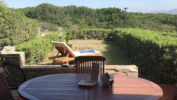 An outdoor patio with a round wooden table and chairs, surrounded by greenery and a view of a hilly landscape. - Casa Ibiscus 1 Bedrooms Apartment in Stintino (Stintino)
