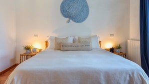 Una cama cuidadosamente hecha con una colcha blanca y varios almohadones, incluyendo uno con la etiqueta 'Sweet Dreams'. Hay dos mesitas de noche con lámparas y pequeñas macetas. Un tapiz azul texturizado de gran tamaño está centrado sobre la cama.