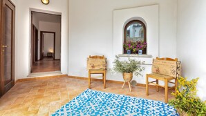 Un pasillo con alfombra estampada, dos sillas de madera, una mesita con una planta y una ventana con flores.