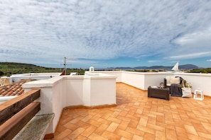 A rooftop terrace with terracotta tiles, a white wall, and a view of distant hills and a sailboat.