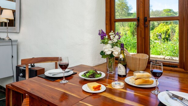 Une table à manger dressée avec des assiettes, des verres et une bouteille de vin, donnant sur un paysage verdoyant par la fenêtre.