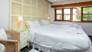 Une chambre avec un grand lit, une table de chevet, un fauteuil en osier et une fenêtre donnant sur un paysage verdoyant.