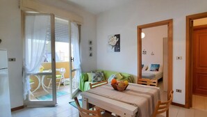 Um espaço de estar compacto com área de jantar, um sofá verde, uma pequena mesa com uma fruteira e uma porta de vidro deslizante para uma varanda.