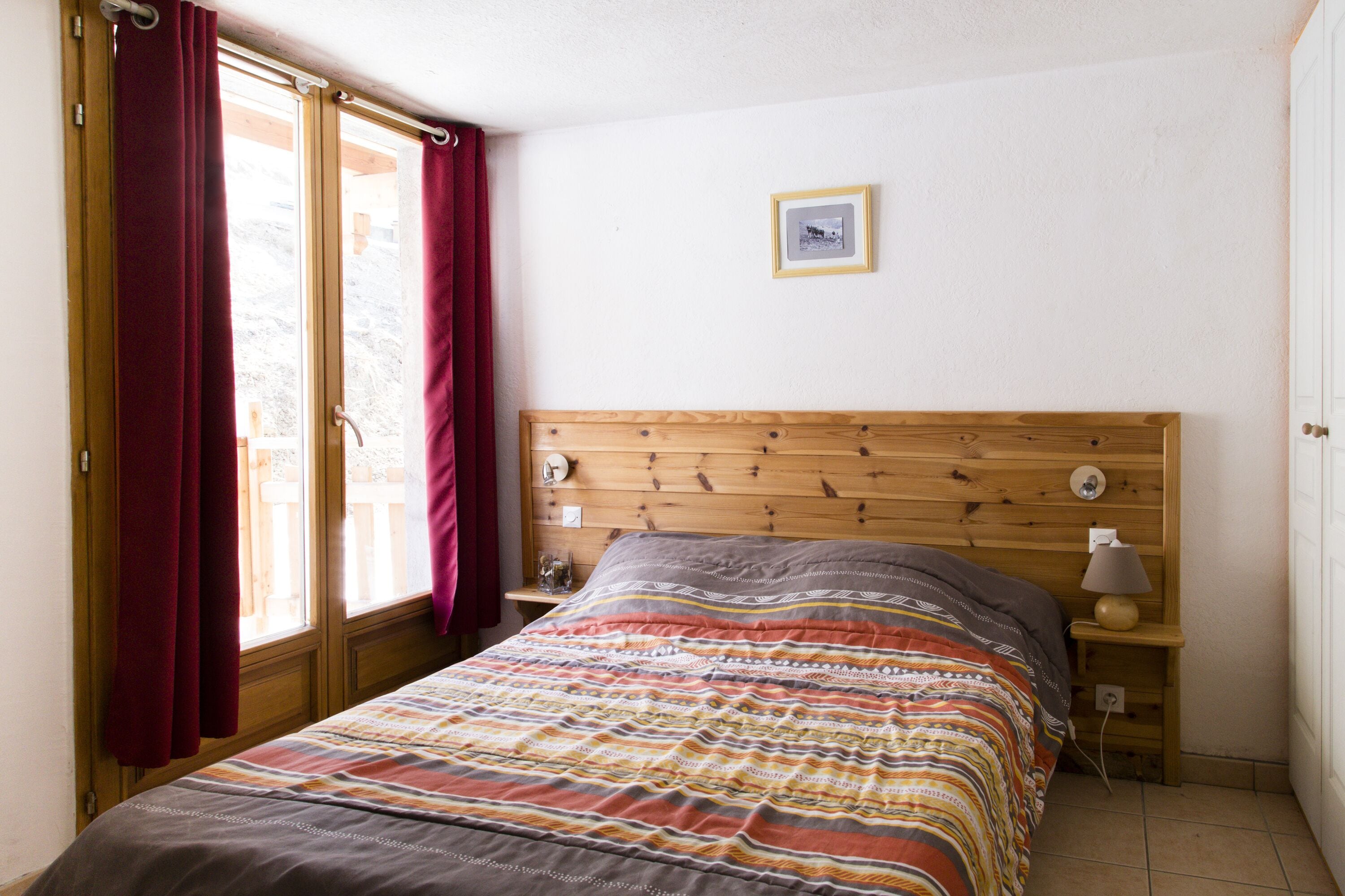 Un dormitorio con cabecera de madera, edredón a rayas de colores, mesita de noche con lámpara y ventana con cortinas rojas.