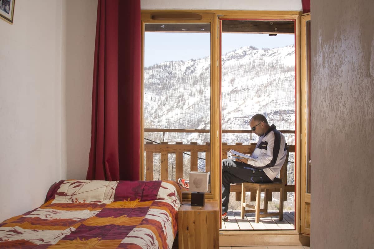 Une chambre avec un lit, une fenêtre donnant sur des montagnes enneigées, et une personne assise à une table près de la fenêtre, en train de lire.