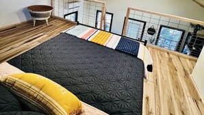 A room with a sloped ceiling, wooden beams, a bed with patterned bedding, a small table, and a view through a window with a grid.