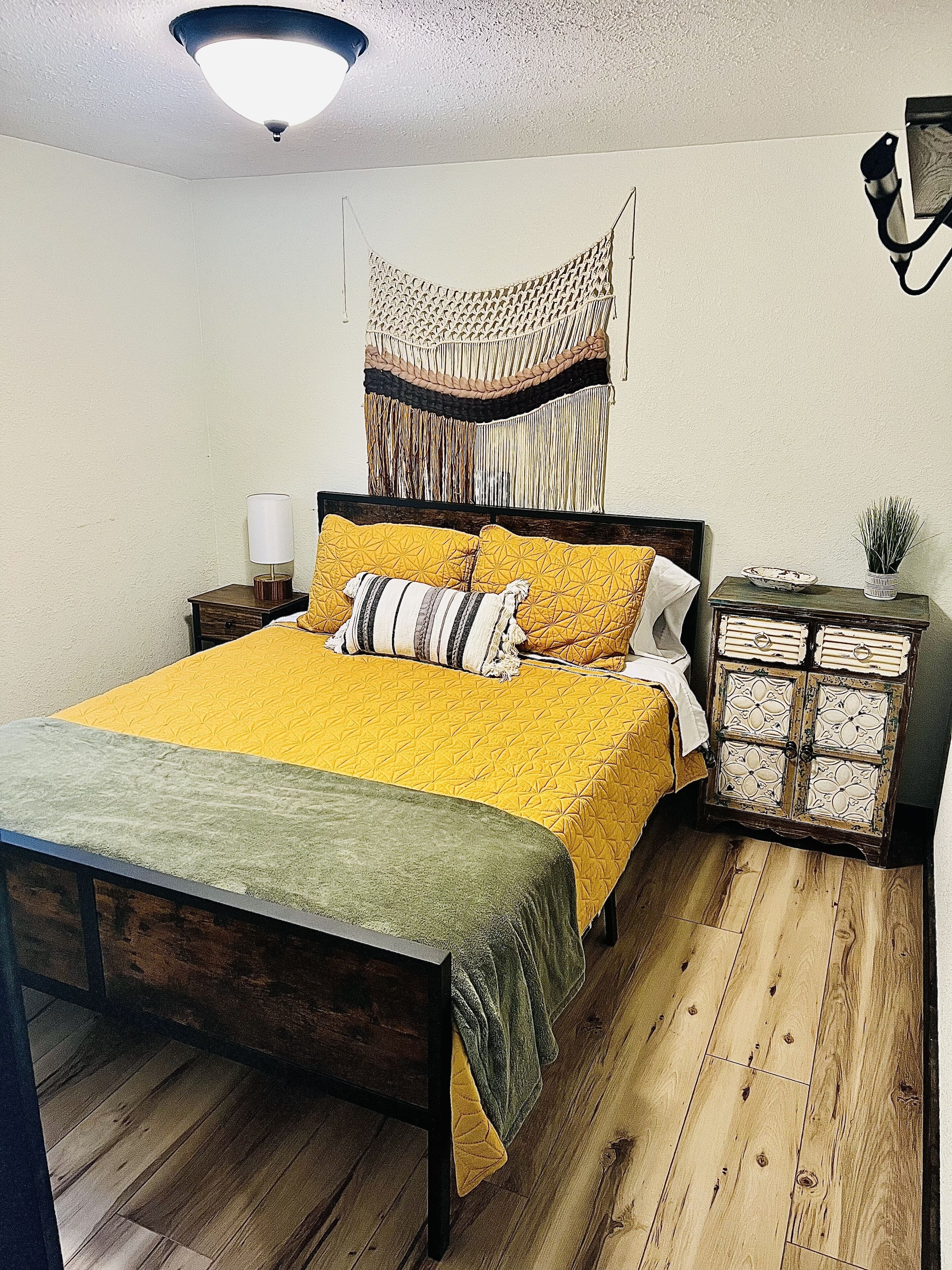 A bedroom with a wooden bed, a bedside table with a lamp, a decorative wall hanging, and a small cabinet with books.