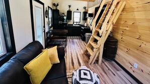 A cozy living room with a black leather sofa, wooden flooring, a wooden staircase, and a kitchen area with a stove and cabinets.
