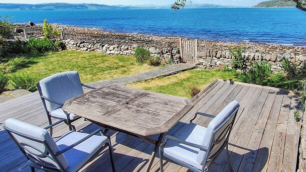 Una terraza de madera con dos sillas blancas y una mesa, con vista a un litoral rocoso y al mar.