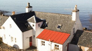 Un edificio blanco con tejado rojo y dos chimeneas, con vistas a una masa de agua tranquila y montañas al fondo.