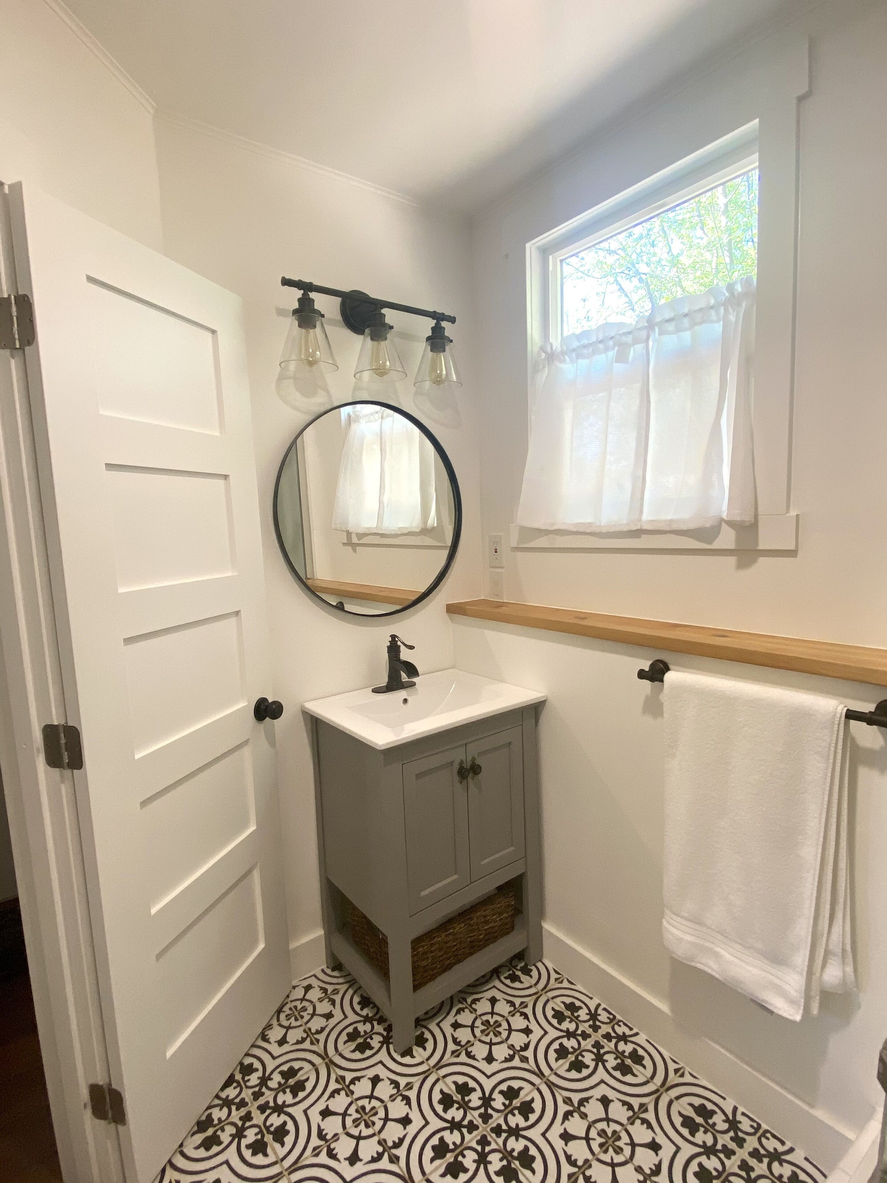 A bathroom with a white door, a sink with a mirror above it, a window with a white curtain, and patterned floor tiles.