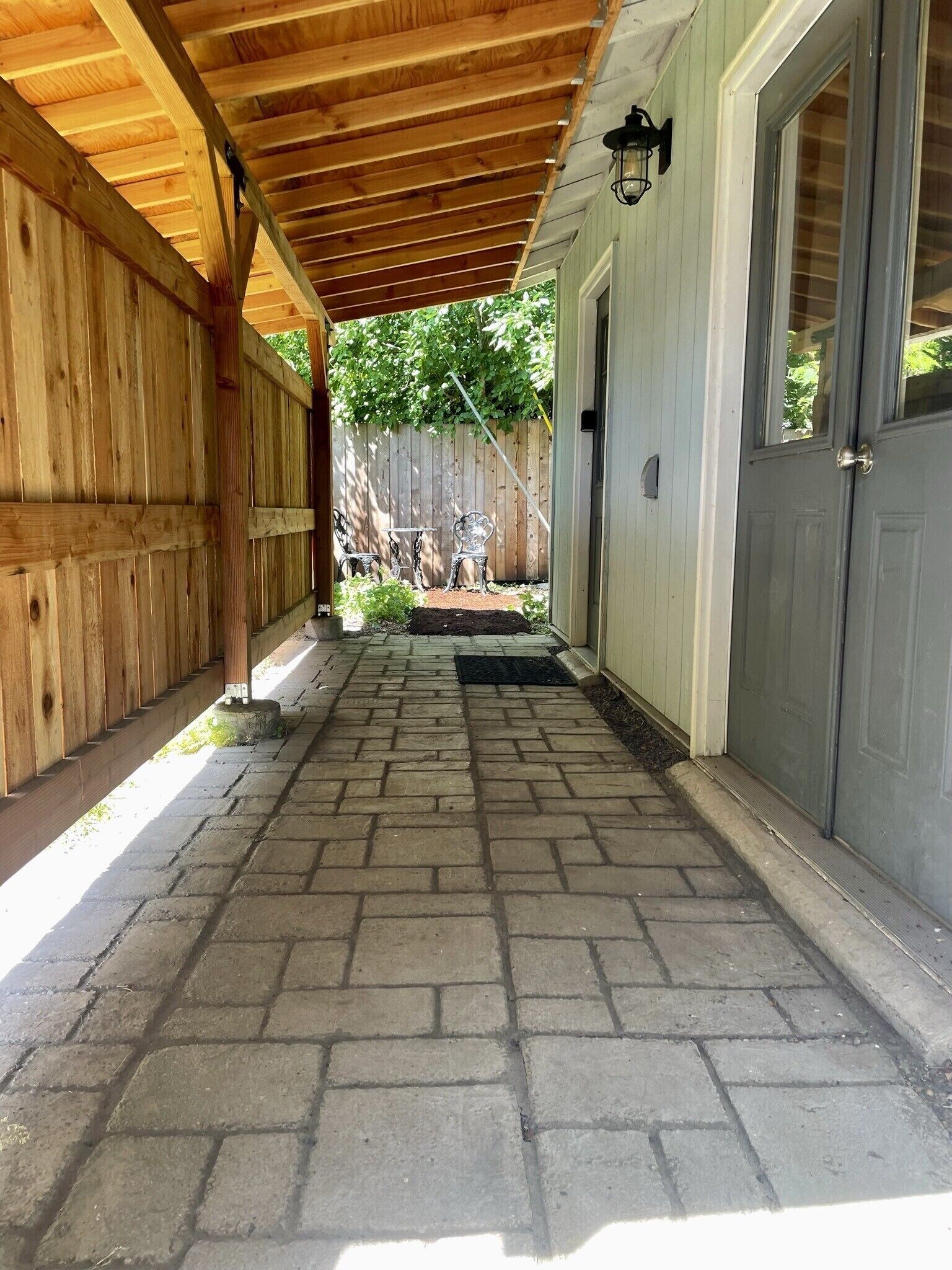 A covered walkway leading to a house with a wooden fence, a door, and a small garden area.