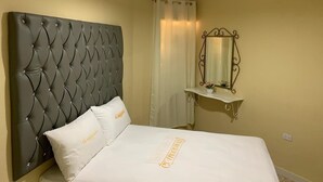 Un dormitorio con cabecera capitonada, cama con ropa de cama blanca, ventilador de techo, espejo y mesita de noche con un florero.