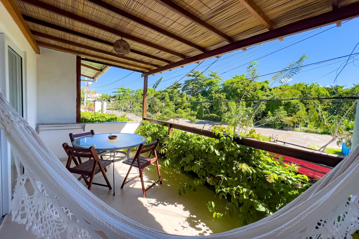 Eine überdachte Terrasse mit rundem Tisch und Stühlen, umgeben von üppiger Bepflanzung und einer Hängematte.