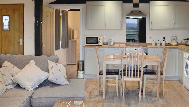 A modern kitchen with white cabinets, a wooden dining table, and a grey sofa with decorative pillows. - 2 bedroom accommodation in Alkham, near Dover (Alkham, near Dover)