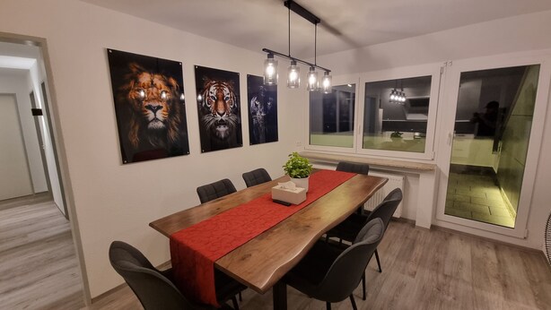 Un coin repas avec une table en bois, un chemin de table rouge et quatre chaises. Trois portraits d’animaux encadrés ornent le mur.