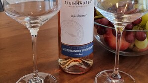 Une bouteille de vin Steinhauser Kressbronner, deux verres à vin vides et un bol de fruits sur une table en bois.