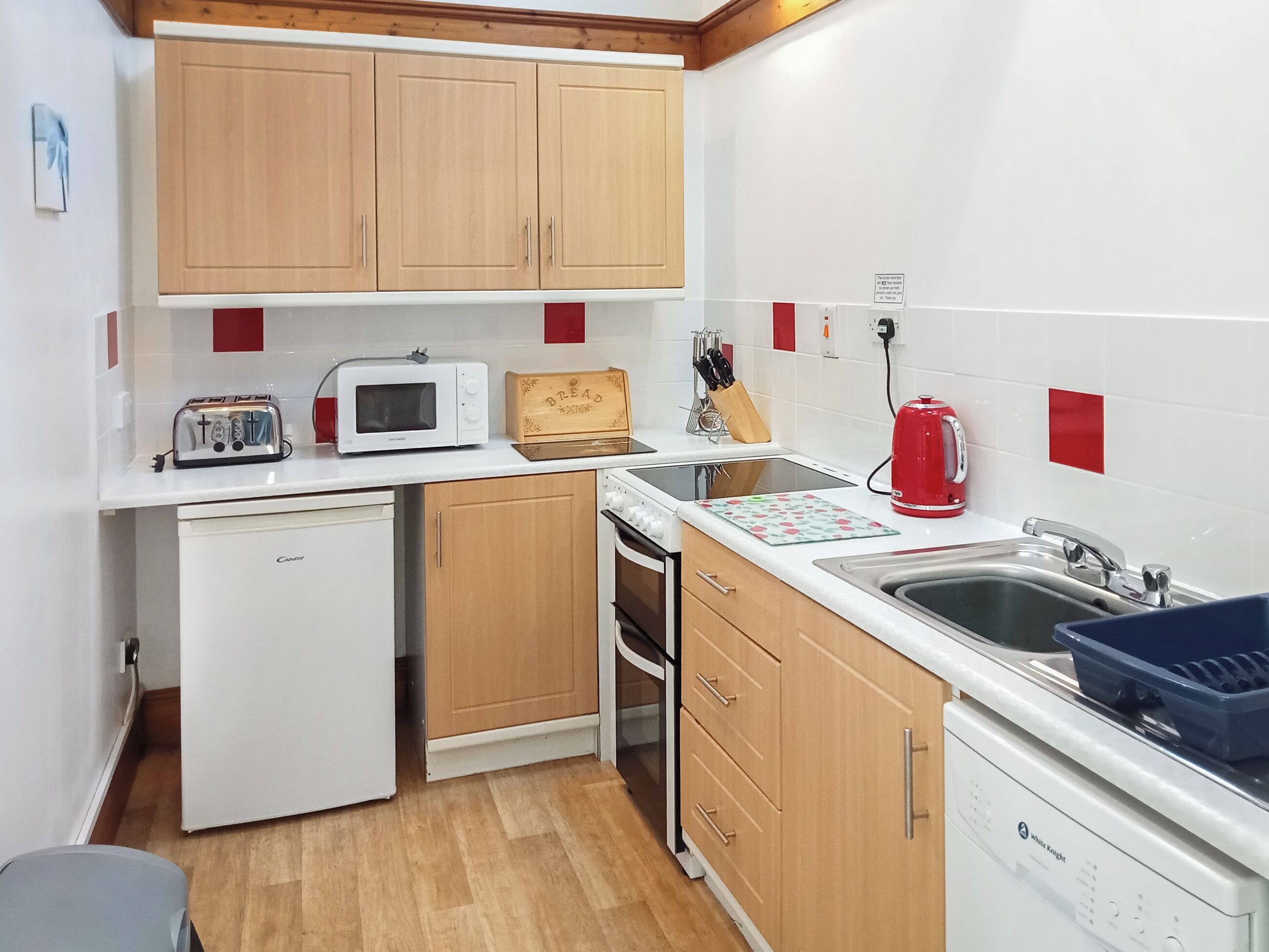 A compact kitchen with wooden cabinets, a white dishwasher, a microwave, a toaster, a red kettle, and a sink with a faucet.