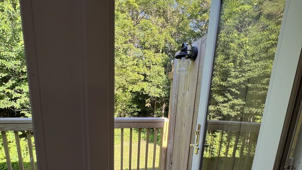 Eine Holzterrasse mit einer Glastür, die zu einem Garten mit Holzzaun und Bäumen führt.