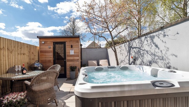 Un jacuzzi, sillas de mimbre, una mesa con una botella y vasos, y una estructura de madera con una puerta.