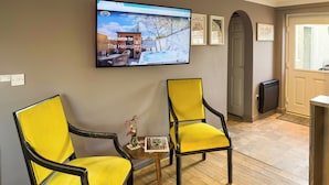 Una habitación con dos sillas amarillas, una mesita con un florero y un folleto, un televisor en la pared y suelo de madera.