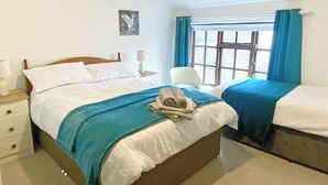 Una cama doble con una manta azul, una mesita de noche con una lámpara, una silla y una ventana con cortinas.