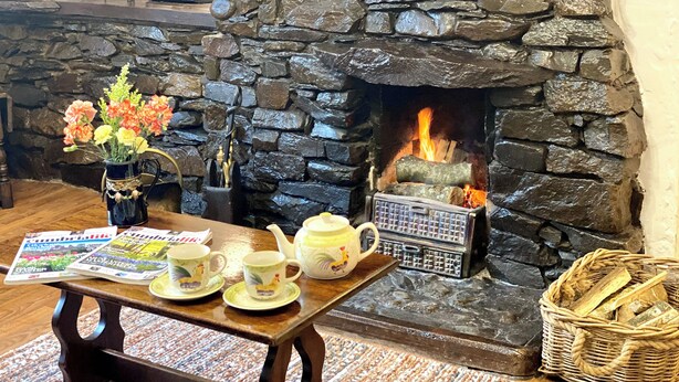 Una chimenea de piedra con fuego, una mesa de madera con periódicos, tetera y tazas, y una cesta de mimbre con leña.