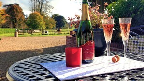 Une bouteille de champagne, deux verres, un bouchon et une bougie rouge sont posés sur une table à l’extérieur, avec en arrière-plan des arbres et une clôture.