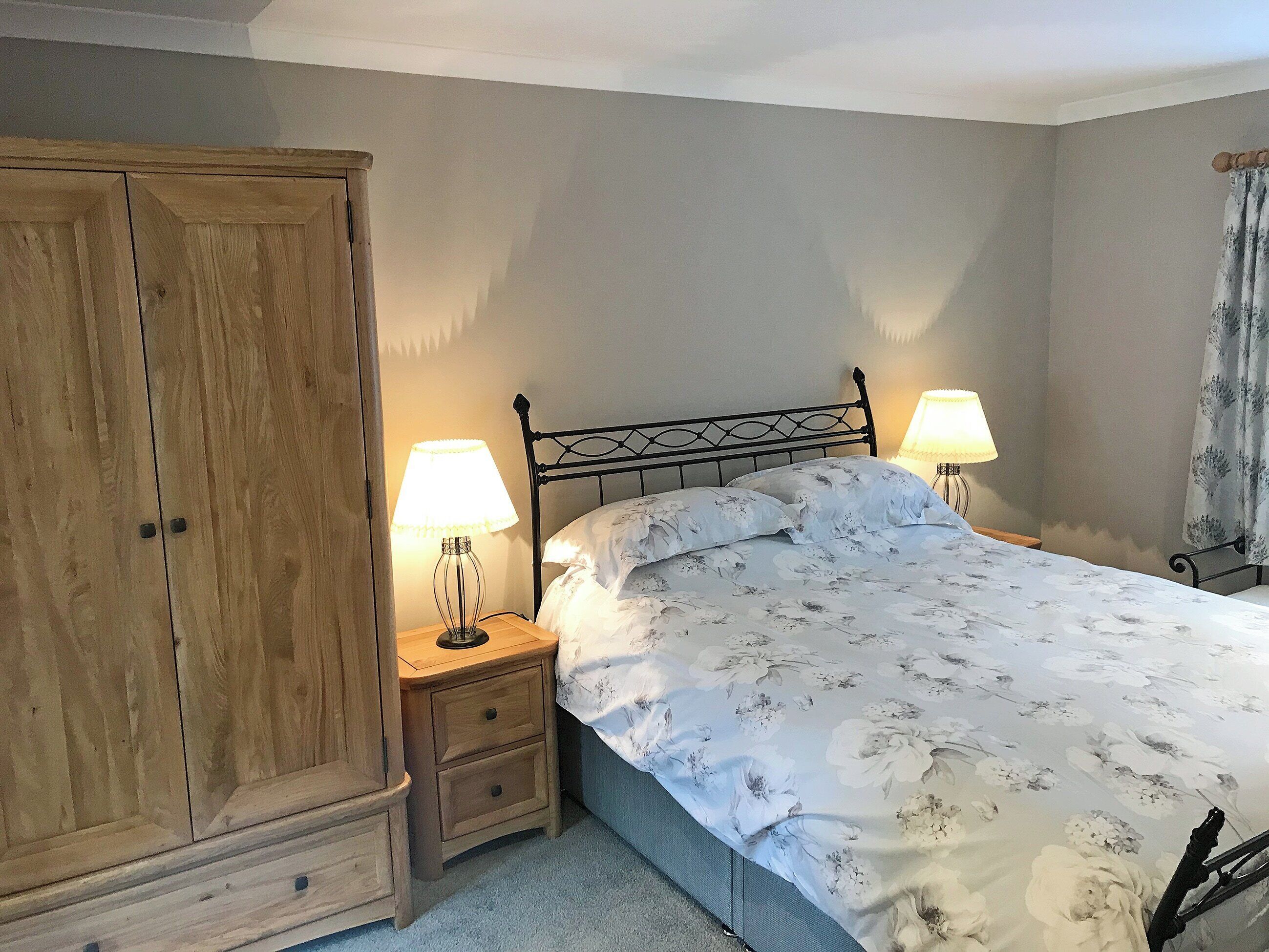 Une chambre à coucher avec un lit, deux tables de chevet avec des lampes, une armoire en bois et une fenêtre avec des rideaux.