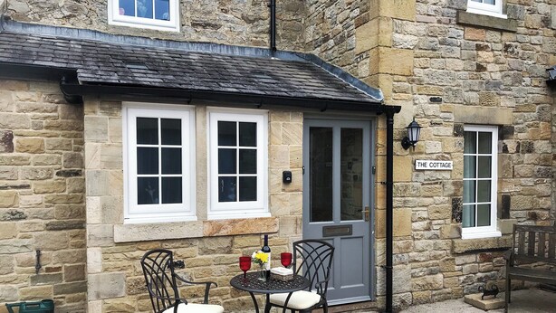 Une petite maison en pierre avec une porte grise, deux fenêtres, une petite table d’extérieur avec des chaises et un paillasson.