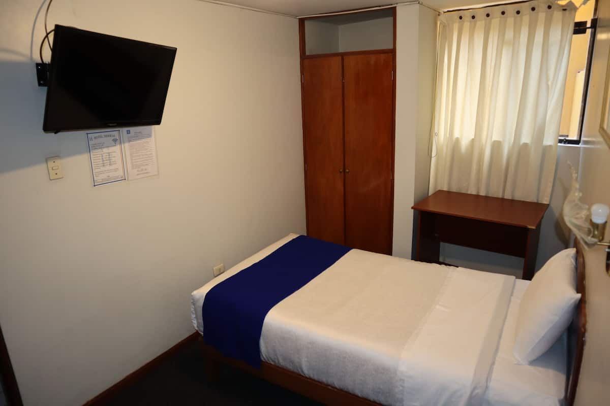Une chambre d’hôtel avec un lit, une armoire en bois, une petite table, un téléviseur fixé au mur et une fenêtre avec des rideaux.