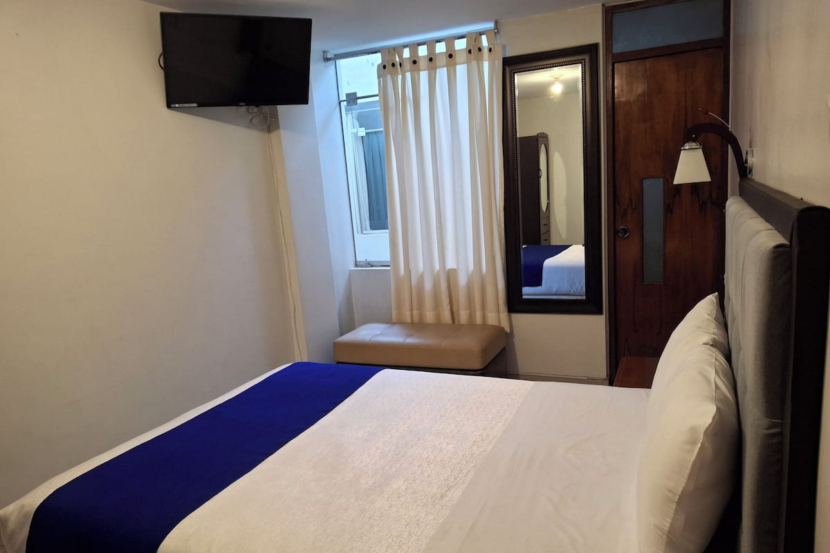 Une chambre d’hôtel avec un lit, un téléviseur fixé au mur, une fenêtre avec des rideaux, une porte en bois et une table de chevet.