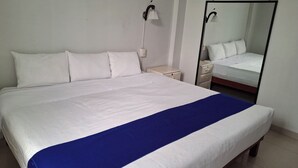 Una cama bien hecha con ropa blanca y un detalle azul, una mesita de noche con una lámpara y un espejo en la pared.