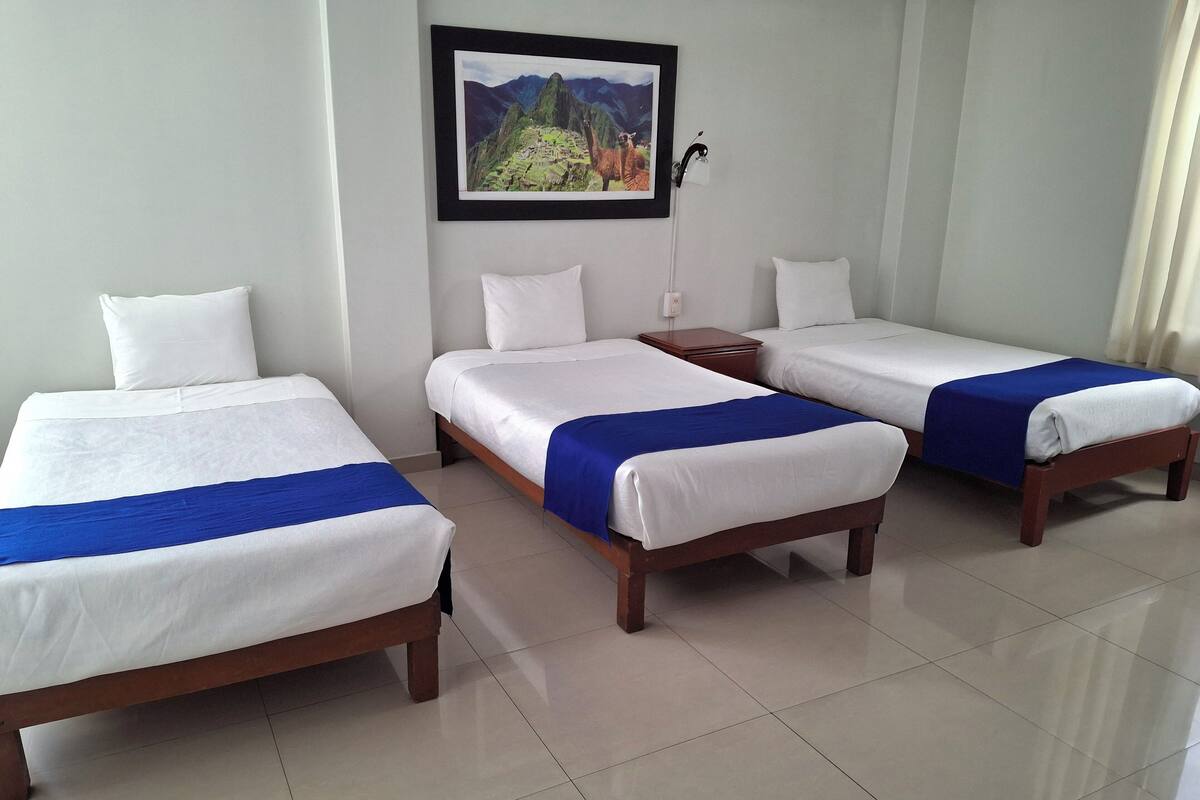 Une chambre d’hôtel avec trois lits simples, chacun agrémenté de draps blancs et d’un élément décoratif bleu. Un tableau encadré représentant le Machu Picchu est accroché au mur, et on aperçoit une table de chevet avec une lampe.
